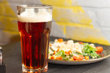 Glass of dark beer with foam and Caesar salad in clay plate on black wooden table