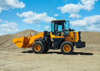 Black-yellow front loader with small wheels against the background of a large pile of stone sand and a blue sky with white clouds. Side view.