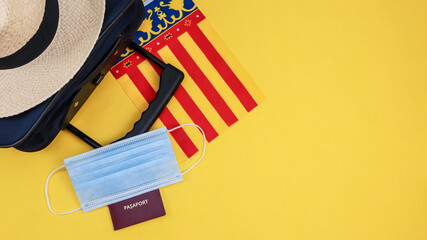 Suitcase and passport with mask.
Suitcase with straw hat, Spanish flag and mask with passport on the left on yellow background with space for text on the right, top view close-up.