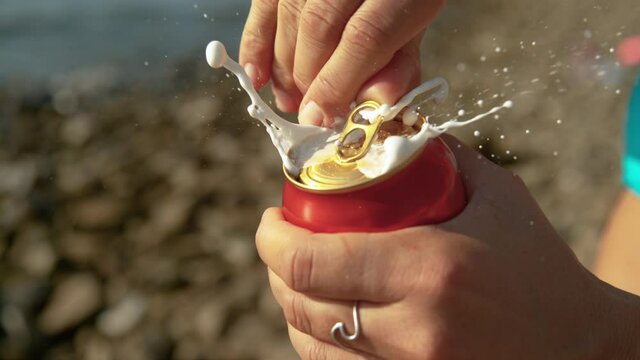 SLOW MOTION, MACRO, DOF: Unrecognizable woman relaxing on the beach opens up a shaken can of beer and gets sprayed with cold foam. Detailed shot of female tourist's hands opening a stirred beer can.