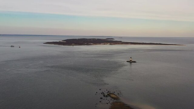 an aerial view of Long Island Sound with a little lighthouse off the east end of Orient Point, NY. It is a cloudy evening at sunset. The drone camera dolly in towards the little lighthouse.