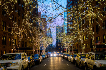 New york city street and brownstone buildings with trees covered in Christmas white lights one point perspective car dusk or early morning