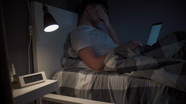 Conceptual illustration of a modern nightmare. Insomnia. A tired, sleepy young man lies in bed and works on a laptop. Eye problems concept. Deadline, overtime. Technology, internet and people