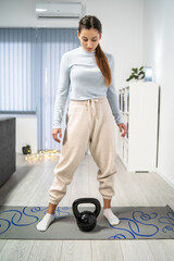 Front view of young caucasian woman training at home - female adult using kettlebell weights in her apartment