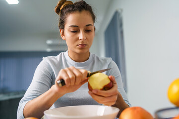 Caucasian young woman peeling fruit while sitting by the table at home - side view on pretty girl holding knife and apple preparing meal - healthy eating concept copy space