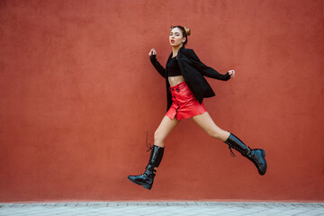 Fototapeta na wymiar Spanish woman, with red mini skirt, jumping, looking at the camera, with a red wall of the street on the background. Spain, young women and fashion concept. Copyspace