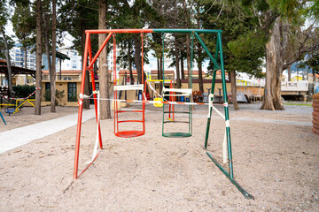 Faulty playground with swings in park is closed for repairs and fenced with red and white signal tape. Prohibition of visiting public places during state quarantine.