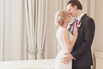 Pretty young couple in elegant wedding clothes tenderly hugging each other in royal room of hotel. Wedding morning of the bride and groom in luxury classic interior. Wedding day ceremony. Young family