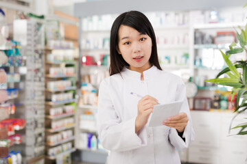 Chinese female looking medicine with notebook near shelves in pharmacy. High quality photo