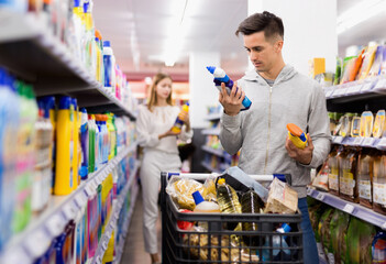 Young glad positive male customer making purchases in supermarket, buying household chemicals - obrazy, fototapety, plakaty
