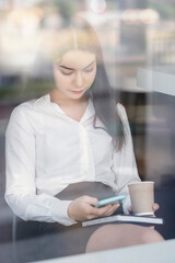 Portrait of young businesswoman using smartphone while sitting at office, looking through glass window.