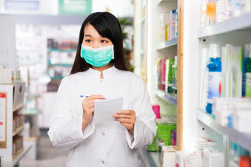 Diligent positive chinese woman pharmacist in protective facial mask keeps track of drugs in interior of pharmacy