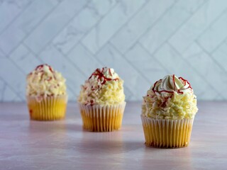 White cupcakes with white frosting swirled high and curled white chocolate with red raspberry drizzle over the top, delicious sweet dessert with pink marble counter and a herringbone tile wall.