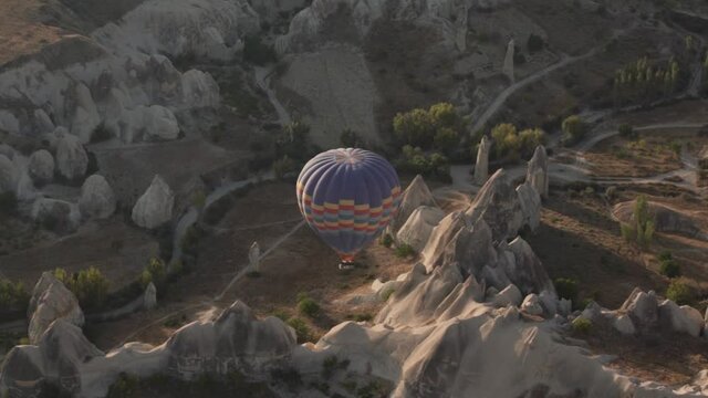 Hot air balloon with striped envelope and basket flies over brown bare rocky hills and stone pillars near ground roads at bright morning sunlight.