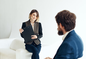 A man in a blue jacket sits opposite a woman in a suit on a light background indoors