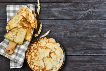 Stacked thin pancakes on a wooden board and plate with a napkin on a wooden background. Traditional wheat pancakes for Shrovetide. View from above. Copy space. Menu.