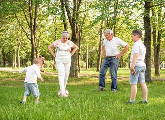 Fototapeta premium Happy family playing soccer on summer meadow. Grandparents with his little cute grandchildren are having fun and playing football on green grassy lawn. Active family play soccer in their leisure time