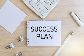 The inscription success plan on a white notebook that lies on a wooden table near a white calculator and a white pen with stationery clips. Business concept