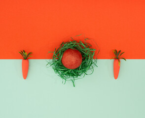 Two carrots and painted easter egg in green paper gras flat lay on green orange background.
