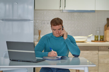 Entrepreneur man on the phone working with a laptop in the kitchen at home. freelancer is negotiating on a smartphone