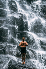 Fototapeta na wymiar A man does yoga at a waterfall. Healthy lifestyle. The concentration of the body. A man does yoga in Bali. A man meditates in nature. Meditation at the waterfall. Young man practicing yoga. Copy space
