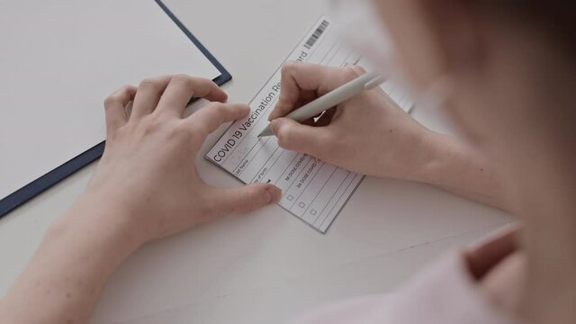 High angle of unrecognizable medical worker filling in Covid-19 Vaccination Record Card with patient name and date of birth
