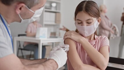Chest-up of cute Caucasian school-aged girl wearing protective face mask, sitting in hospital room, getting painful vaccine injection on shoulder from unrecognizable medical worker