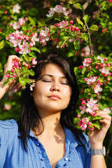 A young attractive plus-size woman with dark hair in a blue dress stands with her eyes closed in a spring blooming garden. Spring time.