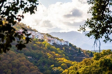 A small old Spanish town is located on a hillside among the autumn forest against a background of mountains and sky with clouds, in the blurred foreground  branches of a tree