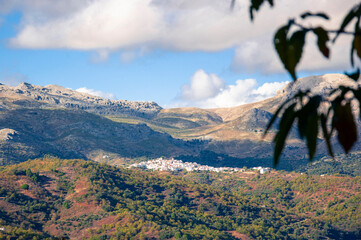 A small Spanish settlement among the mountains, consisting of white houses with red tiled roofs, a bright blue sky with white clouds, shadows from the clouds are falling on the mountains