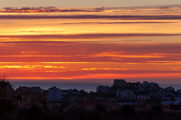 Dusk during the last minutes before sunrise with painted in different shades of red, orange and yellow clouds in the sky over the sea coast with residential buildings.
