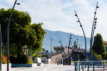 A city park with curved footpaths above the ground on pylons, curved lamp posts, green trees and architectural structures against the backdrop of a mountain range.