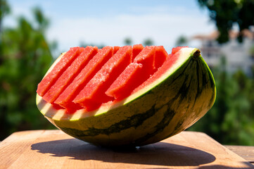 The flesh of a quarter of a watermelon cut into rectangular pieces is on a board in bright sunlight against a blurred background of a garden, building and sky