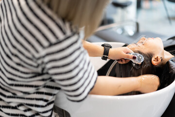 Obraz premium Focus of Asian female customer in plastic face shield to prevent water splashing on eyes and face, laying on hair washing bed having hair washed with water by Asian female hairdresser in hair salon.