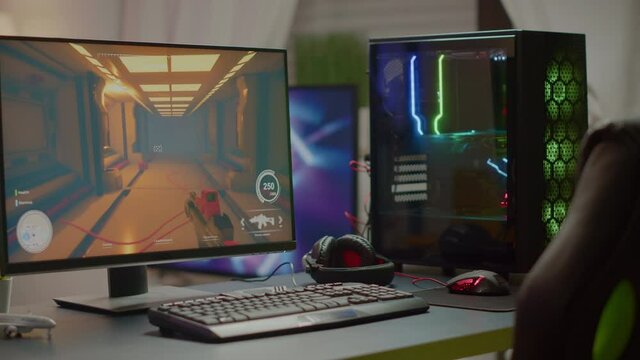 RGB powerful personal computer with first-person shooter game on screen in gaming studio with no people at desk. Empty cozy room with modern design is lit with warm and neon light.
