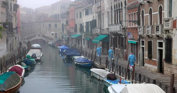 Paramedic people in protective antivirus suits during covid 19 pandemic going to a house near boats moored on canal in city autumnal foggy daytime Venice Italy