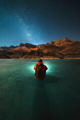 A young man with a backpack sits on the ice of a frozen lake at night in winter and looks at the mountains and the milky way by the light of the moon