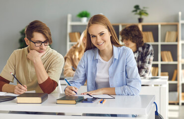 Back to school and offline education concept. Pretty happy smiling caucasian teenage female student writing in notebook and looking at camera. Pupils of highschool or college studying in classroom