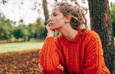 Caucasian young woman resting in the park. Female wearing an orange knitted sweater has pensive...