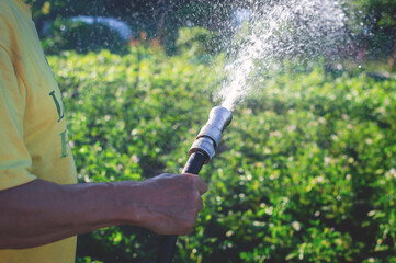 Hand of an senior man with a watering hose, a stream of water pours a lawn in the garden in the day...