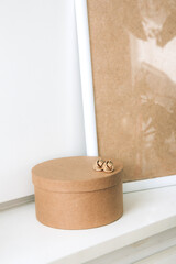 Still life. Blank beige poster frame, craft round box and beautiful gold earrings on the white wall. Side view