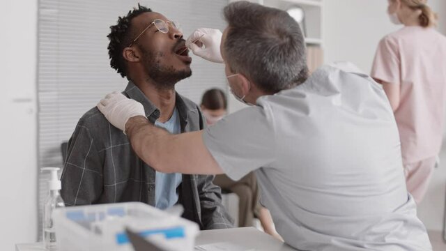 Medium close-up of grey-haired male nurse wearing medical mask, taking DNA sample with cotton swab from mouth of African man, sitting in hospital