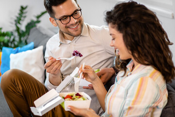 Smiling couple sitting on sofa and eating fast food salad at home.
