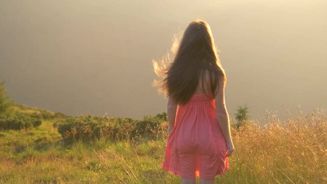Young woman in red dress standing on grassy field on a windy evening in autumn mountains enjoying view of nature.