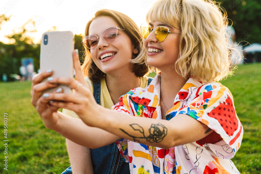 Poster Image of excited two women in sunglasses taking selfie on smartphone