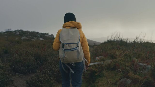 Female solo traveller hike in rough rugged terrain during storm. Wind blow her hair around, woman hiker in yellow puffy jacket with textile backpack. Cinematic travel wanderlust