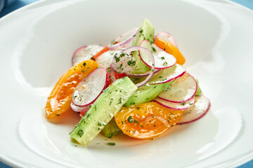 Fresh and light vegetable salad of tomatoes, cucumbers and radishes in a white plate on a blue tablecloth.