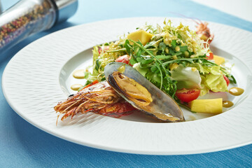 Appetizing salad with seafood, langoustines or shrimp, mussels, arugula and cherry tomatoes in a white plate on a blue tablecloth