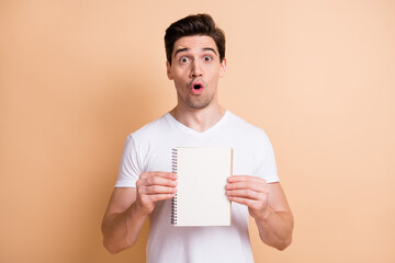 Portrait of impressed brunet man show notebook wear white t-shirt isolated on beige color background