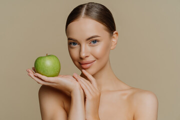 Photo of shirtless brunette young woman with combed hair holds fresh green apple uses natural cosmetic has bare shoulders isolated over brown background. People cosmetology body care concept